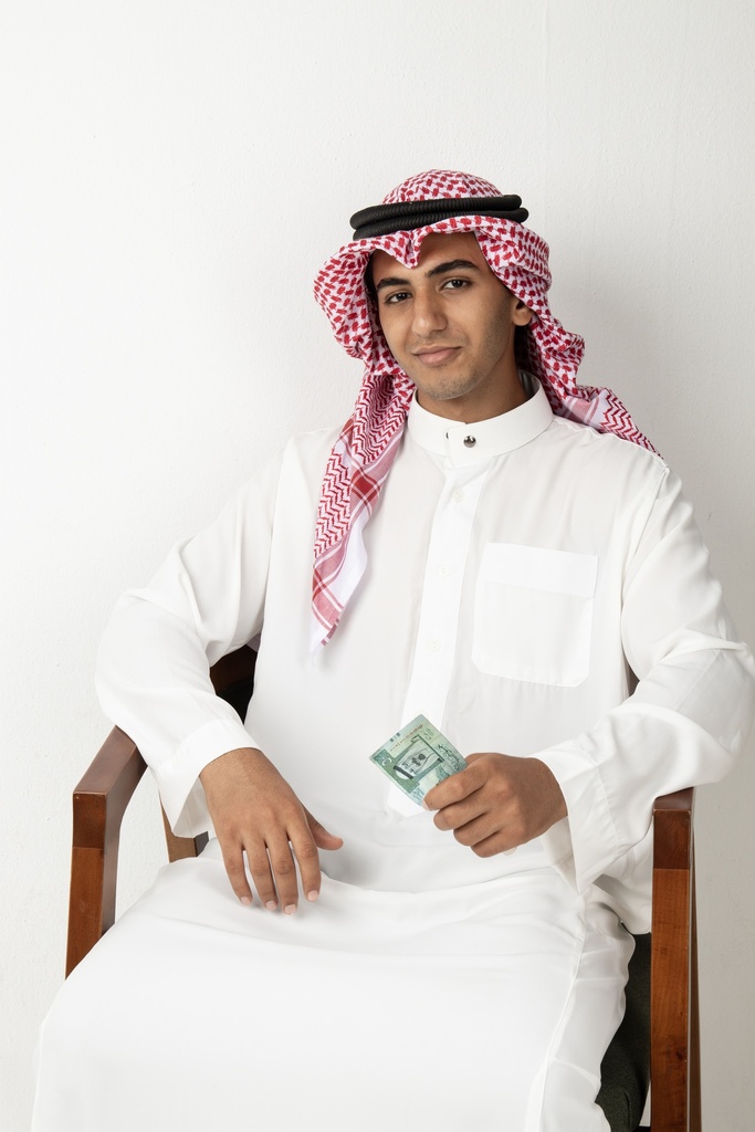 A young Gulf man sitting with a Saudi currency in his hand