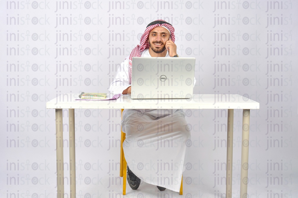 Saudi or khaliji man working on a laptop and talking on the phone in his office.