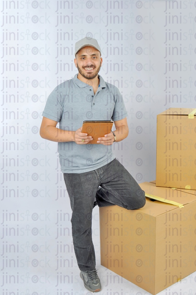 A delivery guy wears a gray cap t-shirt uniform works as dealer courier with the purchasing boxes holding a tablet in his hands