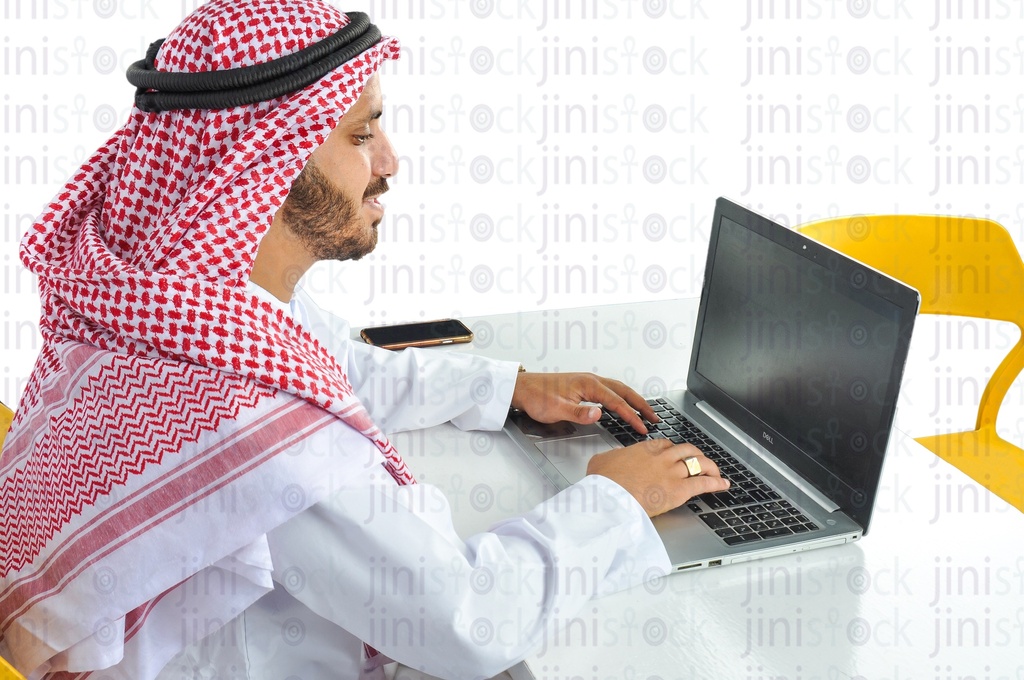 over-shoulder shot of a Saudi, Khaliji,  Emirati or a man from the Gulf working on a laptop in his office.