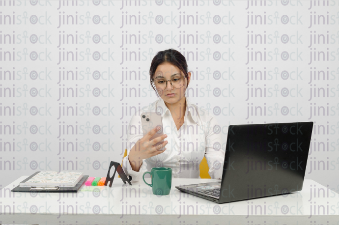 Woman sitting at a desk, wearing glasses, holding a mobile phone in her hand - close-up frontal shot - stock image