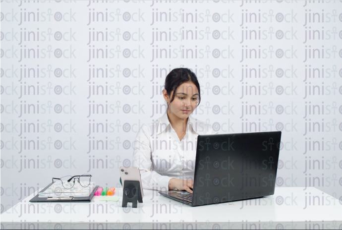 Woman sitting at desk, wearing glasses, working on laptop and smiling - frontal close-up - stock image