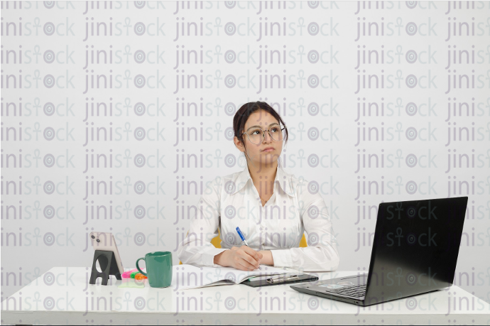 Woman sitting at desk, wearing glasses, writing and looking up - frontal close-up - stock image