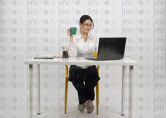 Woman sitting at desk - working on laptop - smiling - wearing glasses - holding a cup of coffee - frontal close-up - stock image