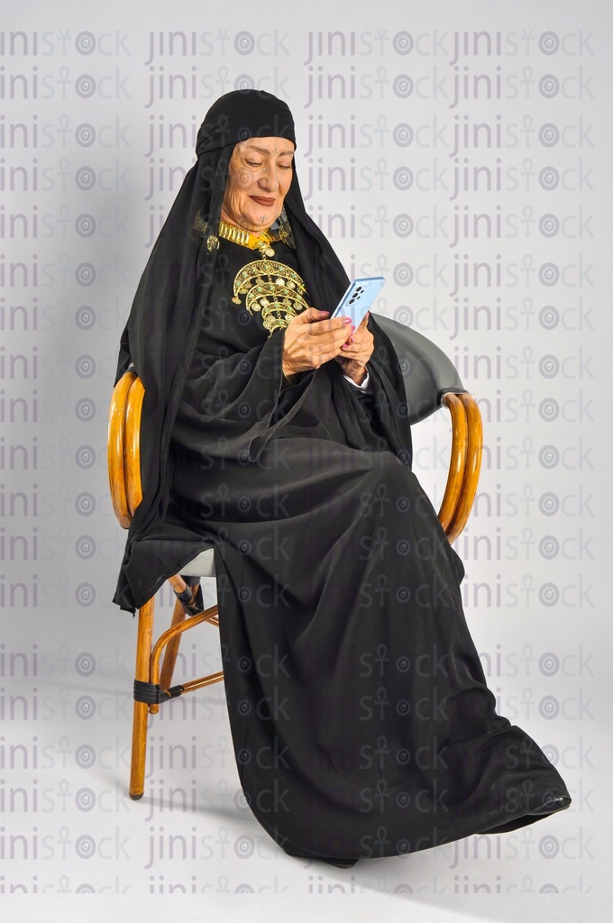 An old upper Egypt woman sitting on a chair and holding a mobile phone and texting on it