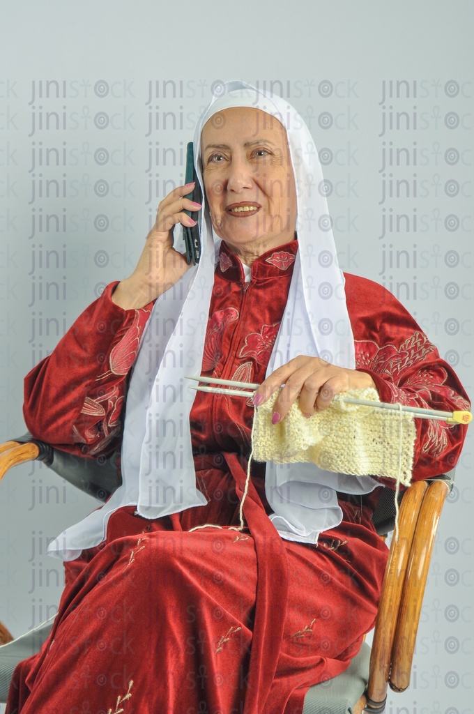 An Egyptian woman sitting on a chair holding a knitting needle and talking on a mobile phone