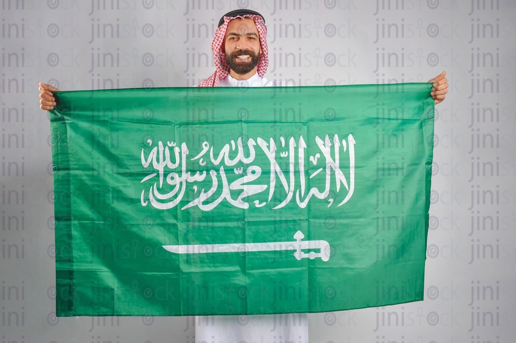 راجل سعودي يبتسم ويحمل العلم السعودي بين يديه احتفالاً بيوم التأسيس السعودي.