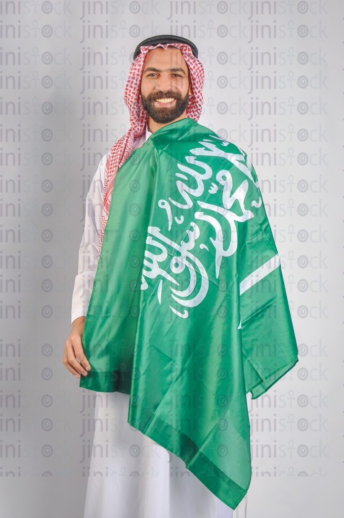 A Saudi man smiling and wearing the Saudi Arabian flag to celebrate the Saudi founding day.