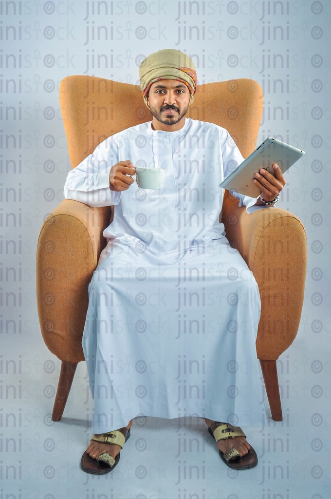 Omani man wearing Omani uniform sitting on a chair and holding a tablet and drinking coffee.