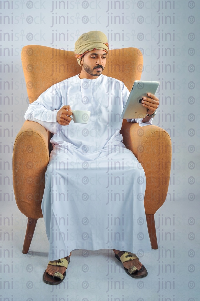 An Omani man wearing Omani uniform working and sitting on a chair looking at a tablet in his hand and drinking coffee.