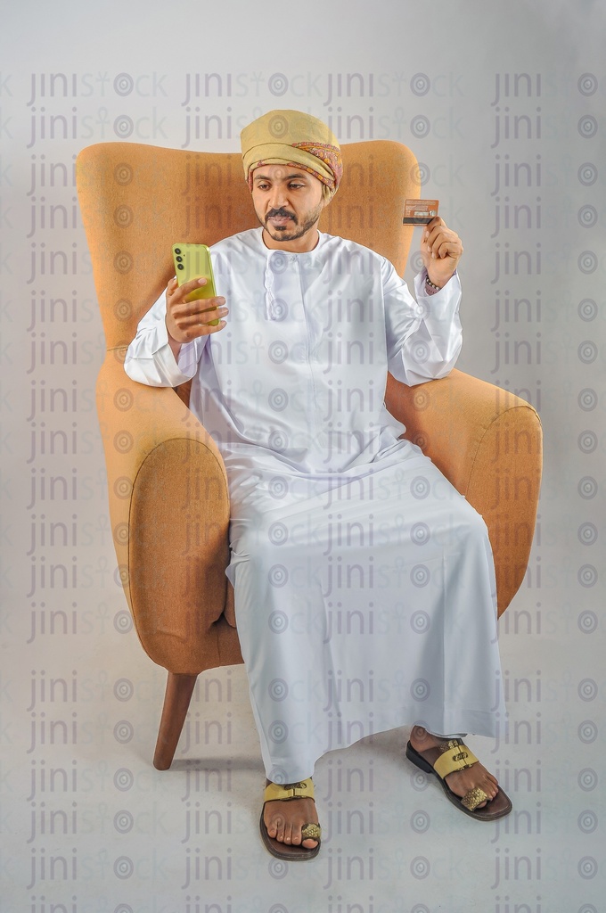 An Omani man wearing Omani uniform and sitting on a chair holding a mobile phone and visa card in his hands.