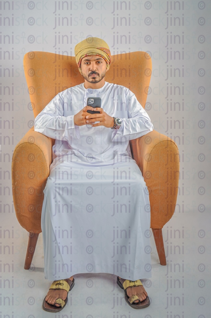 An Omani man wearing Omani clothes sitting on a chair holding a mobile phone browsing the offers and looking at the camera