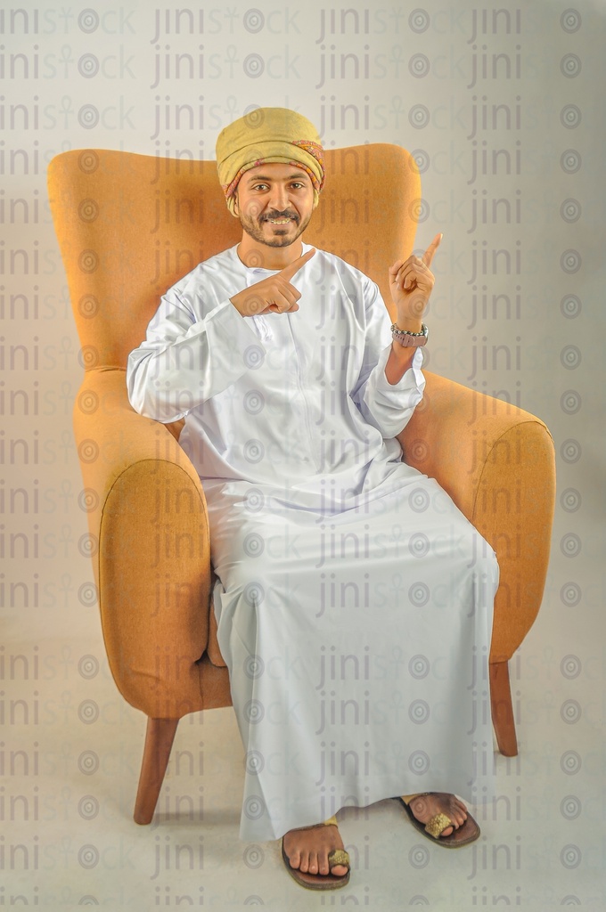 An Omani man wearing Omani clothes sitting on a chair and pointing at something next to him