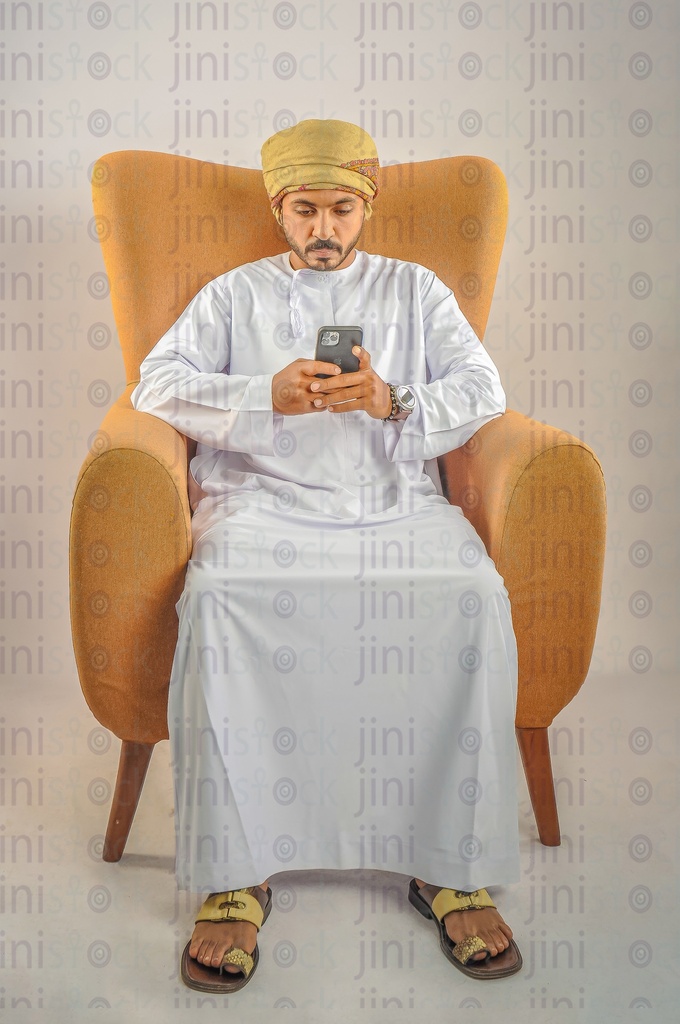 An Omani man wearing Omani clothes sitting on a chair holding a mobile phone and texting or browsing the offers Via mobile applications.