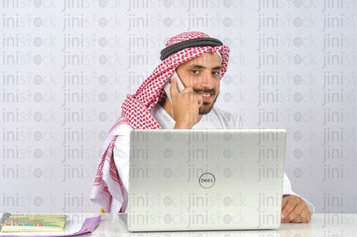 رجل سعودي أو خليجي يعمل على جهاز كمبيوتر محمول ويتحدث عبر الهاتف في مكتبه.