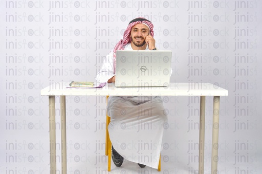 رجل سعودي أو خليجي يعمل على جهاز كمبيوتر محمول ويتحدث عبر الهاتف في مكتبه.