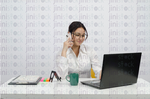 Woman sitting at a desk - working on the laptop - talking on the mobile phone - with a cup of coffee in front of her - close front shot - stock image