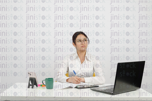 Woman sitting at desk, wearing glasses, writing and looking up - frontal close-up - stock image