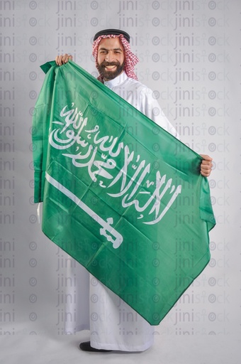 A Saudi man smiling and holding the Saudi Arabian flag in his hands to celebrate the Saudi founding day.