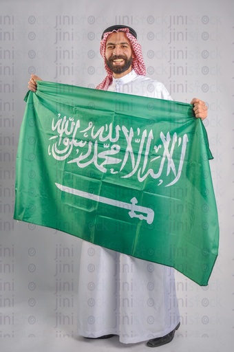 A Saudi man smiling and holding the Saudi Arabian flag with his hands to celebrate the Saudi founding day