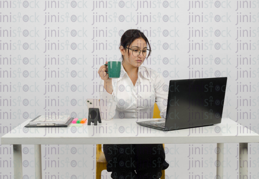 Woman working on a laptop holding a cup of coffee, wearing glasses - close-up front shot - stock image