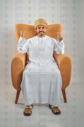 An Omani man wearing Omani clothes sitting on a chair and doing the winner sign