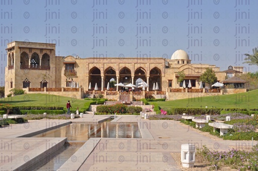 A building in an Islamic style in a garden, while there is a corridor in the middle of the green spaces for the passage of people.
