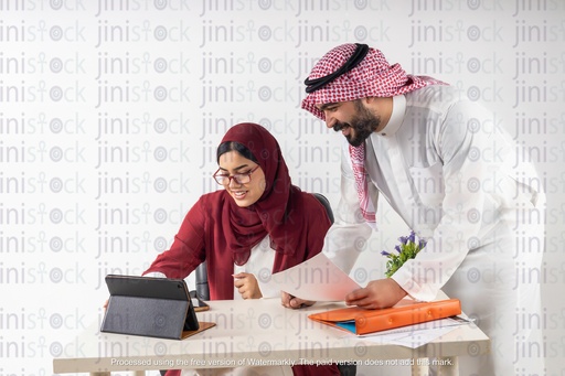 saudi woman khaliji sitting behind a desk and a man from gulf leaning to check something on the ipad