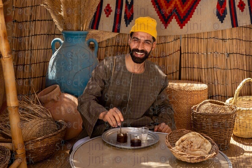 An Egyptian farmer in a wicker hut. In front of him is a tray with tea cups and a cup of sugar 
